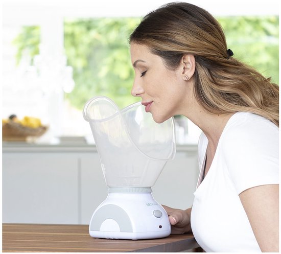 Vrouw met een witte top inhaleert stoom uit een Medisana gezichtssauna met brede, transparante kap.