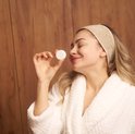 Vrouw met hoofdband en badjas houdt een witte shower steamer omhoog en glimlacht tevreden.