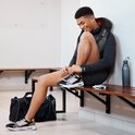 Man in sportkleding zit op een houten bankje, strikt zijn veters, met een zwart/grijs massagekussen op zijn nek.