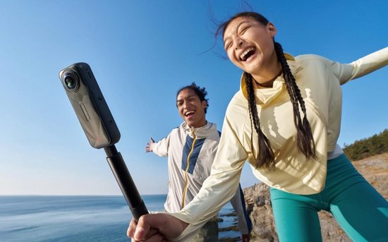 Twee lachende mensen, een man en een vrouw, met Insta360 X5 op een heuveltop bij zee.