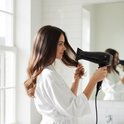 Vrouw met lang bruin haar in witte badjas, droogt haar haar met een zwarte Lafe haardroger.