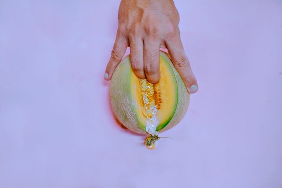 Main sur un melon coupé en deux, avec les graines et le jus qui en dégoulinent sur fond rose.