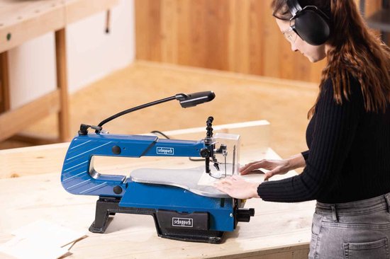 Vrouw met gehoorbescherming gebruikt een blauwe Scheppach figuurzaagmachine in een werkplaats.