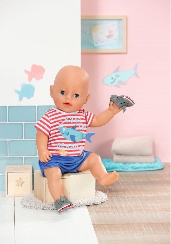 Baby Born pop zittend, met rood-wit gestreept shirt met haai en blauwe broek. Draagt grijze haaienschoentjes. Houdt een haaienschoen vast.