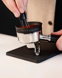 Een portafilter met gemalen koffie en een tamper-tool, staand op een zwarte tampermat.