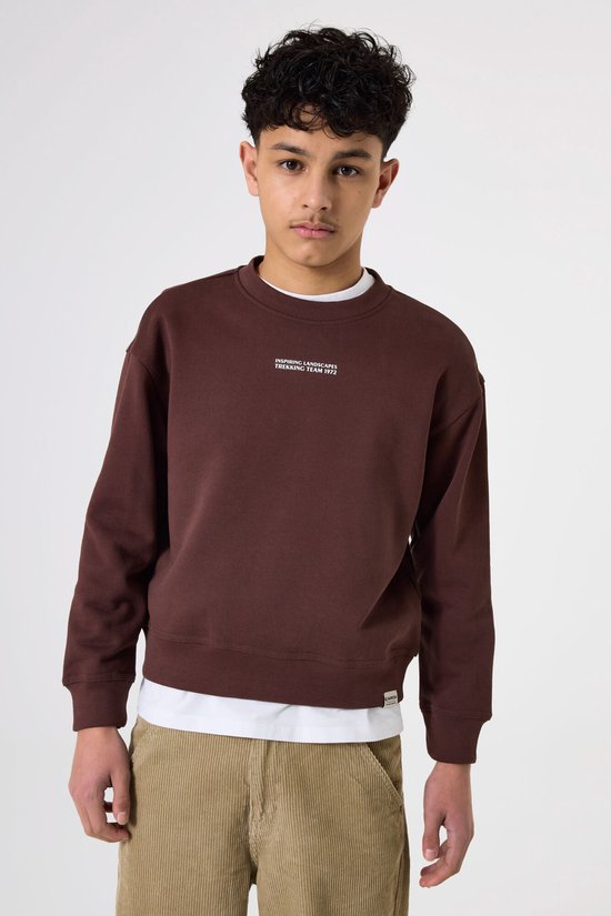 Jongen in bruine sweater met tekst 'INSPIRING LANDSCAPES TREKKING TEAM 1972'.