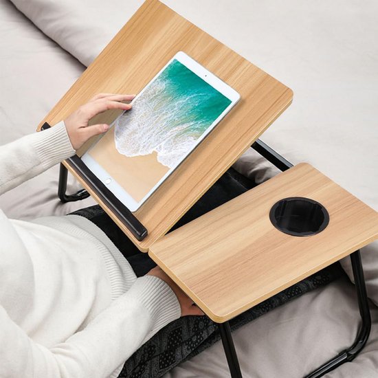Vrouw op bed met een lichtbruine houten laptoptafel op schoot. Tafel met bekerhouder en tablet.