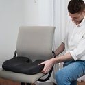 Een man met een wit overhemd plaatst een zwart orthopedisch zitkussen op een grijze bureaustoel.