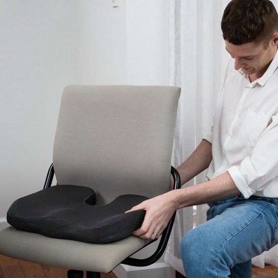 Een man met een wit overhemd plaatst een zwart orthopedisch zitkussen op een grijze bureaustoel.