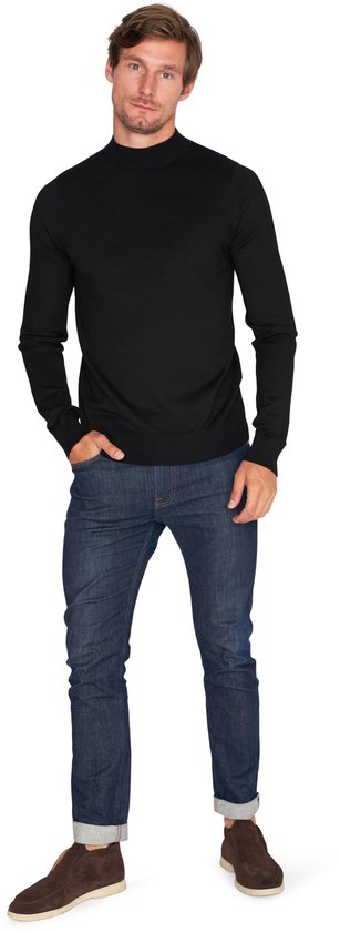 Man staat met handen in zakken, draagt zwarte coltrui en jeans.