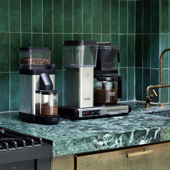 Twee Moccamaster koffiezetapparaten op aanrecht met groene tegels. Eén zilver, één zwart-zilver met bonen en gemalen koffie.
