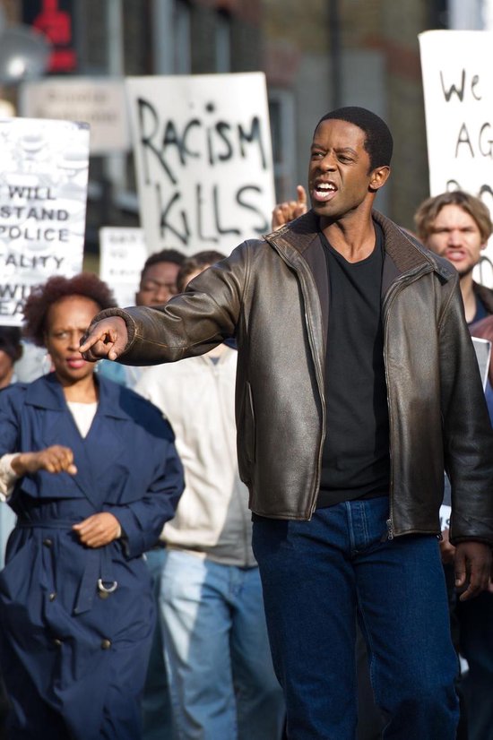 Een donkere man in een bruine leren jas wijst terwijl hij een groep demonstranten leidt. Achter hem een bord met 'RACISM KILLS'.