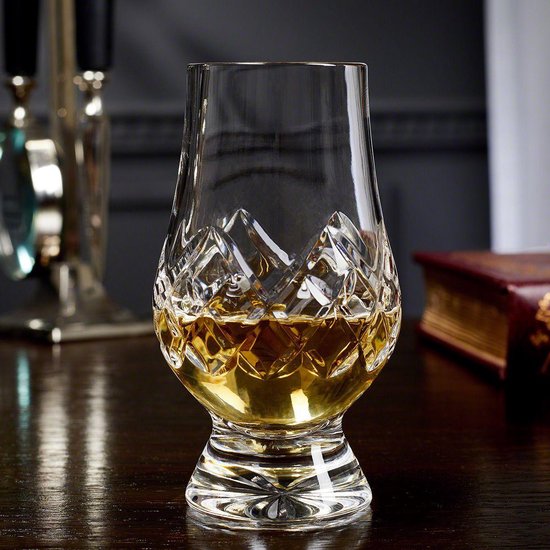 Glencairn whiskyglas met amberkleurige drank, geplaatst op een houten tafel naast een boek.