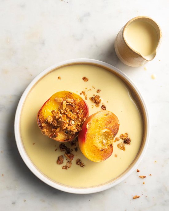 Gegrilde perzik, gehalveerd en bestrooid met granola, geserveerd op een bord met gele saus en een kannetje room.