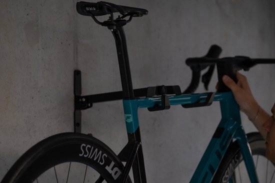 Zwarte racefiets met blauw frame hangt aan zwarte fietsmuurbeugel, een hand past het aan.