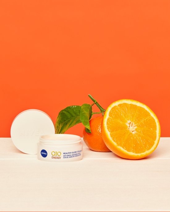 Nivea Q10 Energy gezichtscreme potje met oranje fruit en groene blaadjes, op een witte en oranje achtergrond.