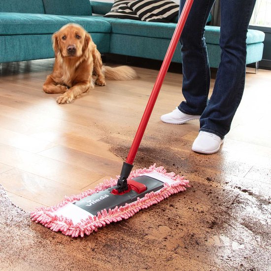 Vileda mop avec manche rouge retirant des poils d'un plancher en bois. Chien couché sur un canapé.