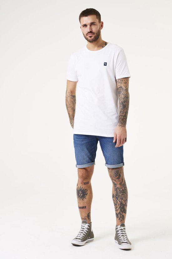 Homme tatoué portant un t-shirt blanc et un short en jean bleu avec des ourlets retroussés.