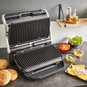 Open contactgrill met grillplaten. Ingrediënten voor burgers, zoals broodjes, augurken en kaas, liggen ernaast.