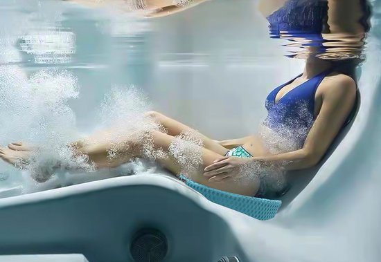 Een vrouw in een blauw badpak zit op een blauw spa zitkussen in een bubbelbad, met water dat rond haar benen stroomt.