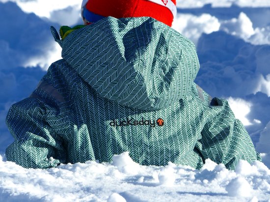 Kind in groen regenpak met visgraatpatroon zit in de sneeuw.