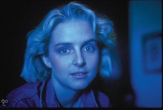 Een vrouw met kort blond haar en blauwe ogen kijkt ernstig in de camera, badend in blauw licht.