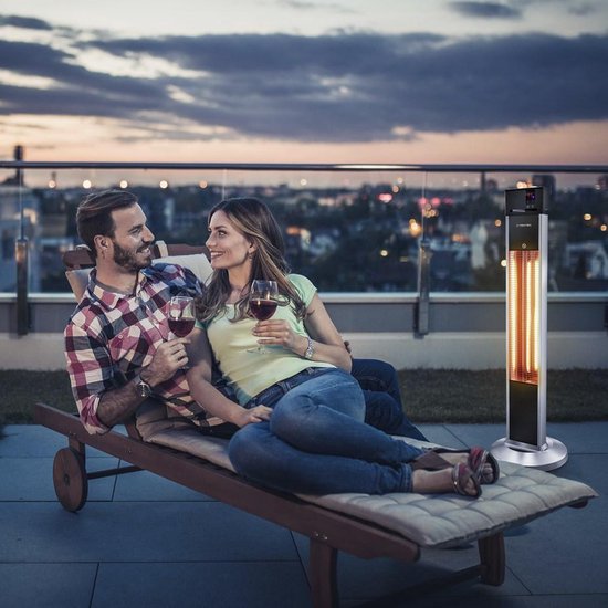 Koppel op loungebank op terras met Trotec terrasverwarmer bij zonsondergang, met stad op achtergrond.
