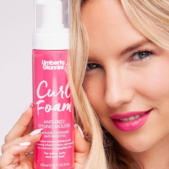 Vrouw met blond haar houdt een roze fles Umberto Giannini Curl Foam omhoog. De fles heeft witte letters 'Curl Foam'.