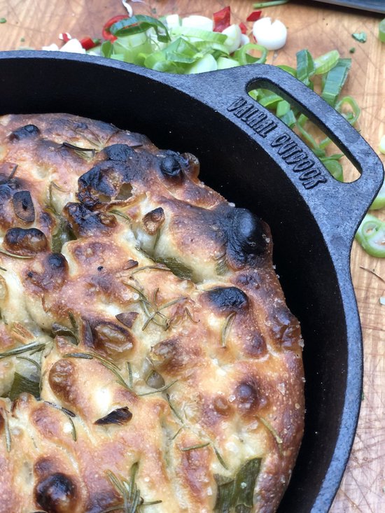 Gietijzeren koekenpan met een goudbruine focaccia, gegarneerd met rozemarijn en rode ui.