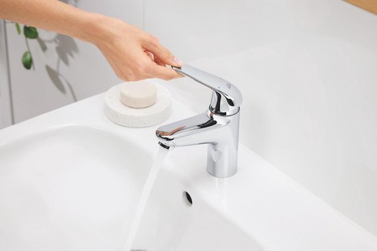 Een hand draait de hendel van een chromen kraan die water geeft op een wastafel.