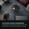 Grijze stofzuiger met een actieradius van 8,5 meter, staand op een donkergrijs vloerkleed bij een marmeren salontafel.