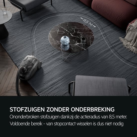 Grijze stofzuiger met een actieradius van 8,5 meter, staand op een donkergrijs vloerkleed bij een marmeren salontafel.