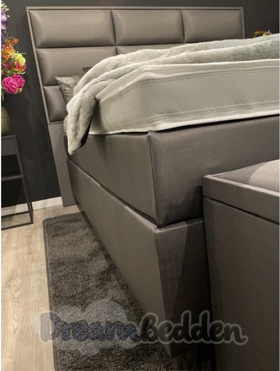 Grijze boxspring met gestoffeerd hoofdbord, witte matras en een grijze opbergbank.