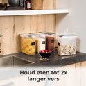 Vier transparante voorraadpotten met pasta, rijst, cacao en ontbijtgranen op een keukenblad naast een gootsteen.