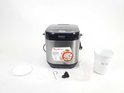 Zilverkleurige broodbakmachine met diverse accessoires, waaronder een maatbeker, binnenpan en deegkneder. Wit aanrecht.