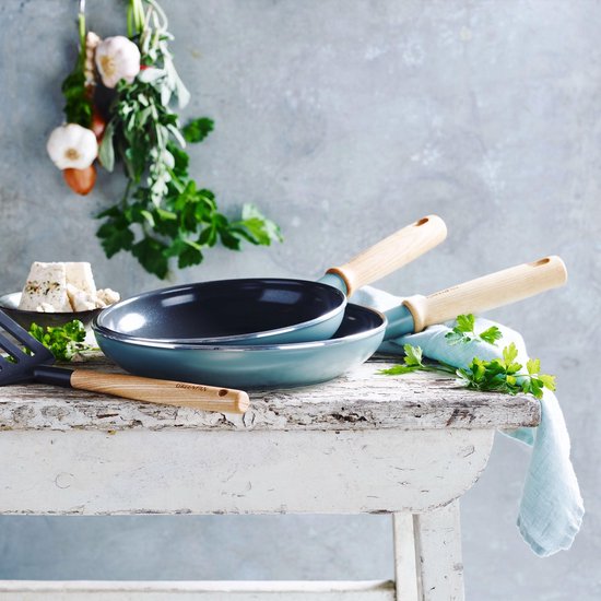 Een blauwgrijze koekenpan met houten handgreep en een tweede pan op een rustieke houten tafel, versierd met kruiden en kaas.