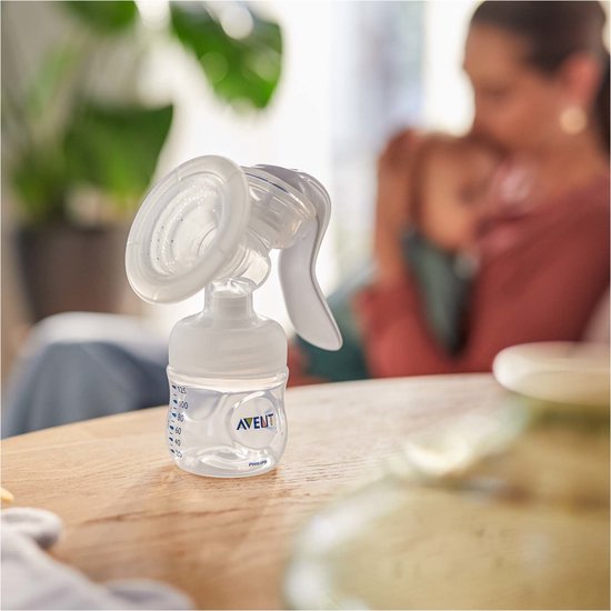 Philips Avent handkolf op tafel. Op de achtergrond een moeder die haar baby vasthoudt. De fles is gegradueerd.