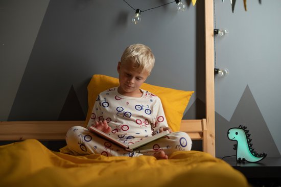 Groen oplichtend dinosaurusspeeltje op een nachtkastje naast een jongen met blond haar die in bed een boek leest.