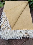 Okergele wollen deken met visgraatpatroon en witte franjes, liggend op een gevlochten bruine tafel.