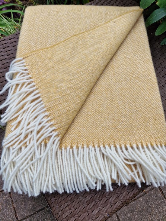 Okergele wollen deken met visgraatpatroon en witte franjes, liggend op een gevlochten bruine tafel.