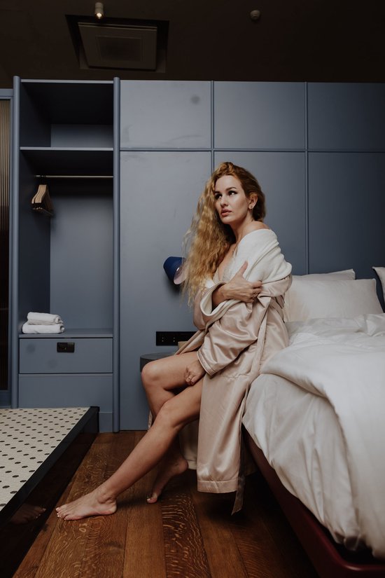 Vrouw met lang blond haar, zittend op een bed in een satijnen badjas.
