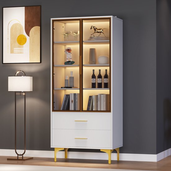 Witte vitrinekast met glazen deuren, gouden handgrepen, gouden poten en warme LED-verlichting. Met decoraties zoals boeken en flessen.
