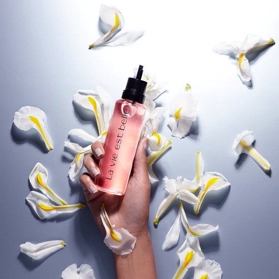 Hand houdt Lancôme La Vie est Belle parfumfles vast, omringd door witte bloemblaadjes. Roze vloeistof.