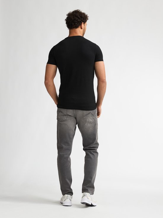 Man met handen in broekzakken, draagt zwarte T-shirt met korte mouwen en grijze jeans.