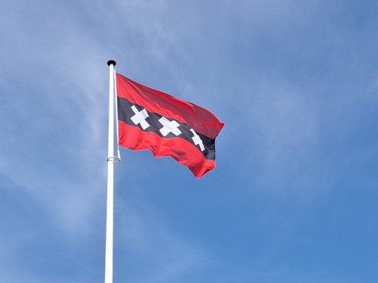 Amsterdamse vlag wapperend aan een witte mast tegen een blauwe lucht.