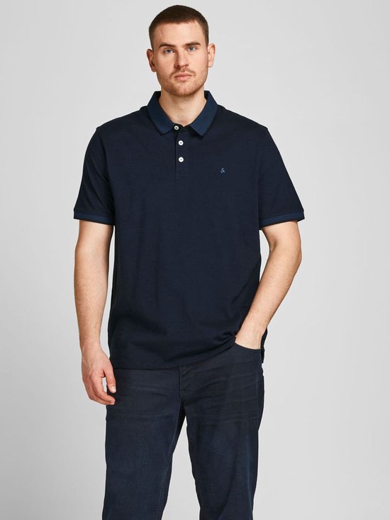 Man in donkerblauw poloshirt met wit geborduurde "&" op borst, navy kraag, drie witte knopen.