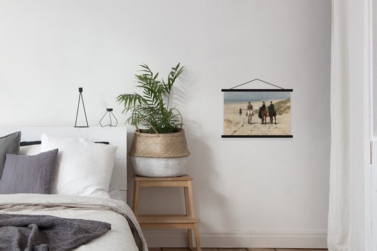 Schilderij met drie mensen te paard op strand, hangend aan een witte muur boven een bed met mand en plant.