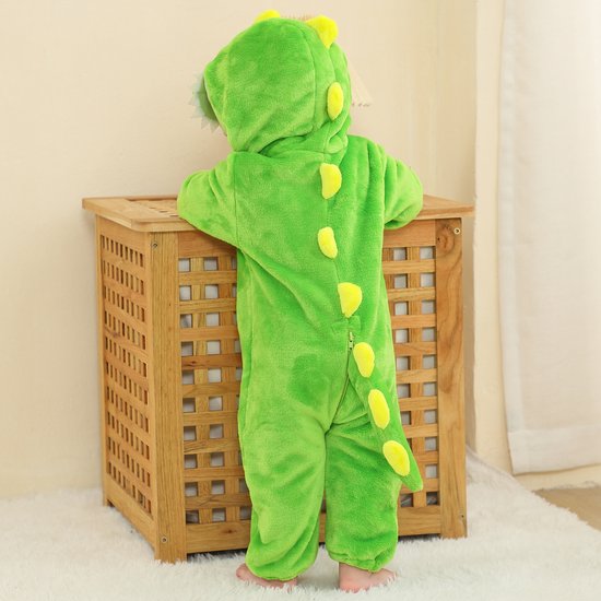 Baby in groen dinosaurus onesie, staand bij houten kast, met staart en rugstekels.