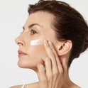 Een vrouw brengt een witte crème aan op haar wang. De crème wordt op de wang gesmeerd.