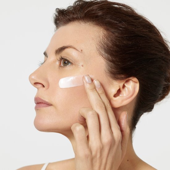 Een vrouw brengt een witte crème aan op haar wang. De crème wordt op de wang gesmeerd.